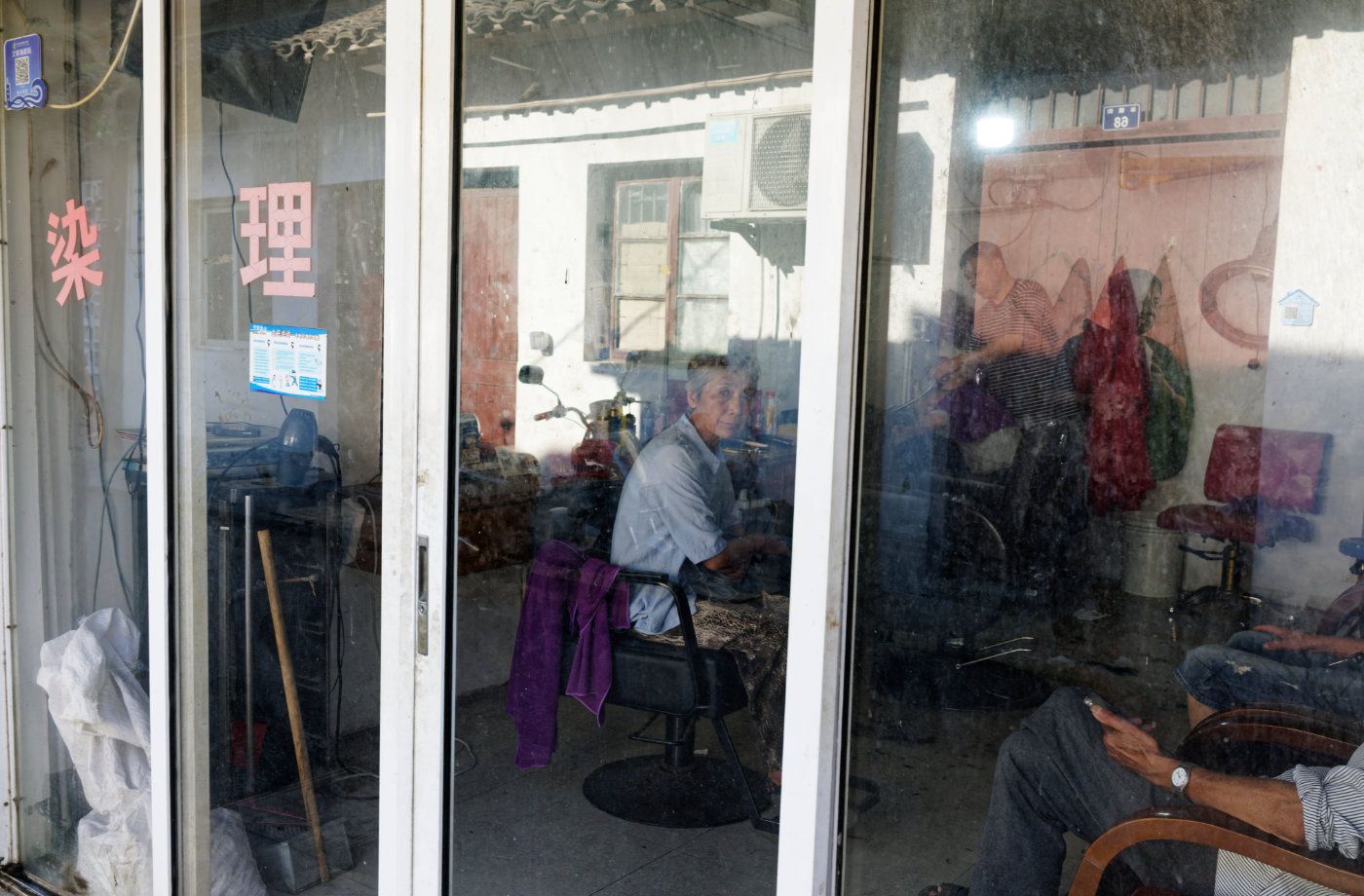 理发店玻璃上同时映出街对面的老屋和屋内等候的人,现实和倒影叠在一起。